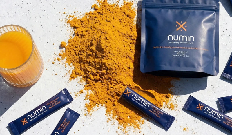 Curcumin ingredient in Numin spilled across a white surface beside the brand's pouch, scattered stick packs, and a glass of orange tea.