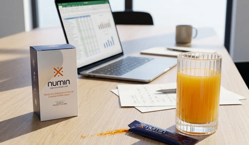 Numin product box and stick pack on a sunlit office desk beside a glass of orange tea, a laptop with spreadsheets, and handwritten notes.