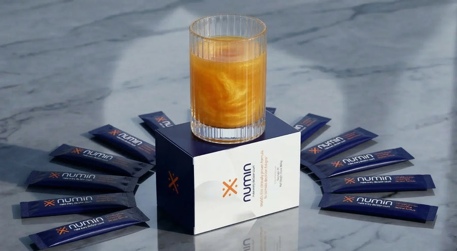Numin drink mix box with a glass of orange beverage and stick packets fanned on marble.