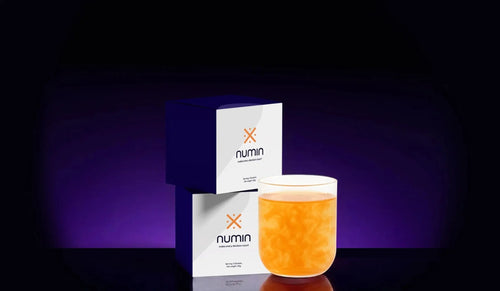 Two Numin supplement boxes beside a glass of orange drink on a purple-lit backdrop.