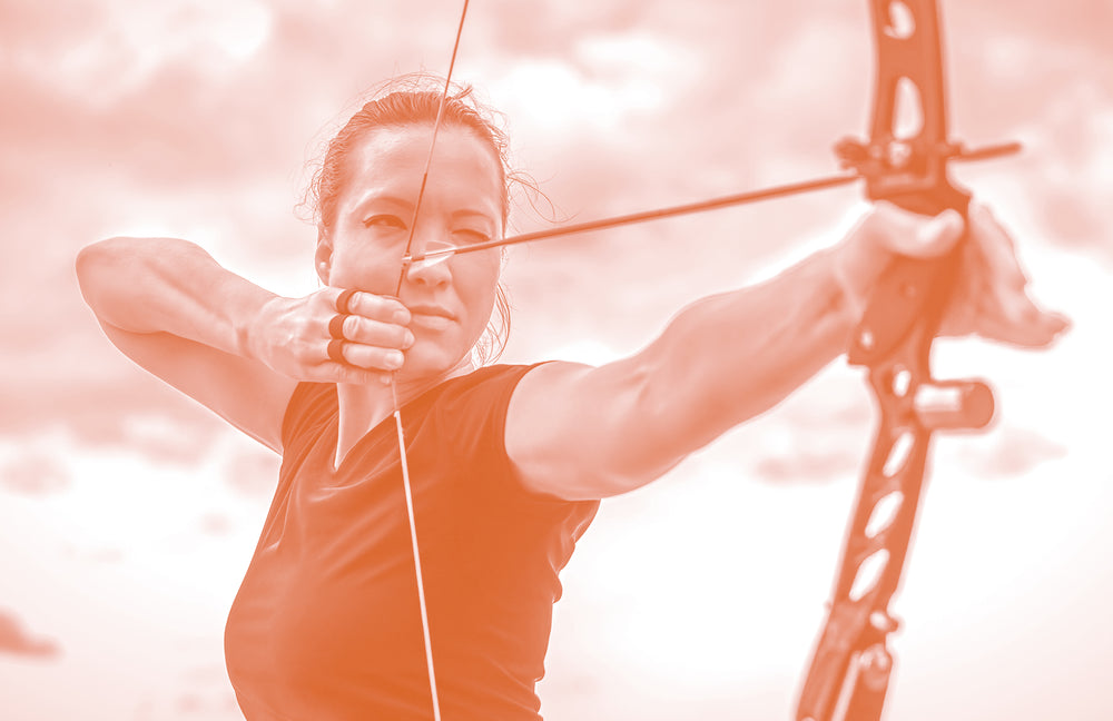 Woman practicing archery with focused concentration, drawing bow and arrow at outdoor target range