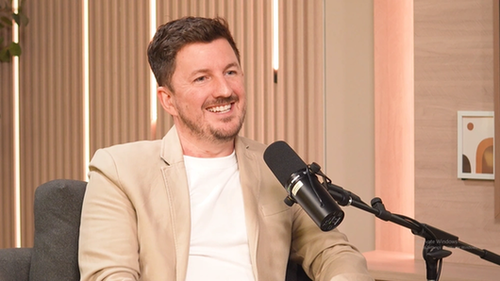 Smiling podcast interview host in beige blazer speaking into microphone in modern studio setting