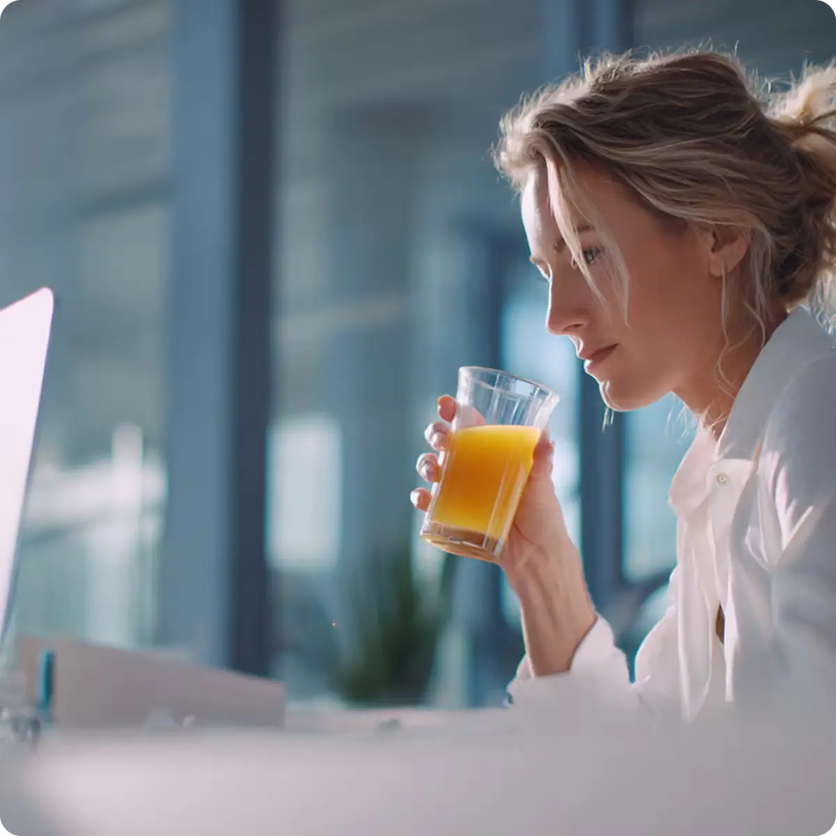 Professional woman in white shirt drinking orange juice while working on laptop at modern office desk